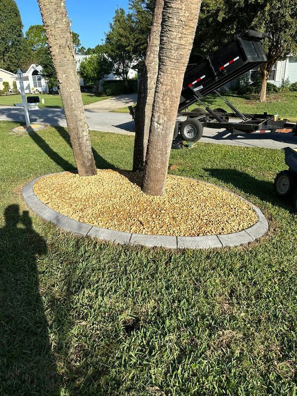 Oval rock bed around palm tree trunks with gray border, on green grass, with a street and vehicle in the background.
