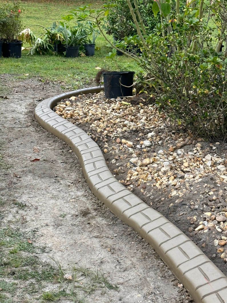 Curving tan brick-patterned border edging a path through a garden with a black bucket in the background.