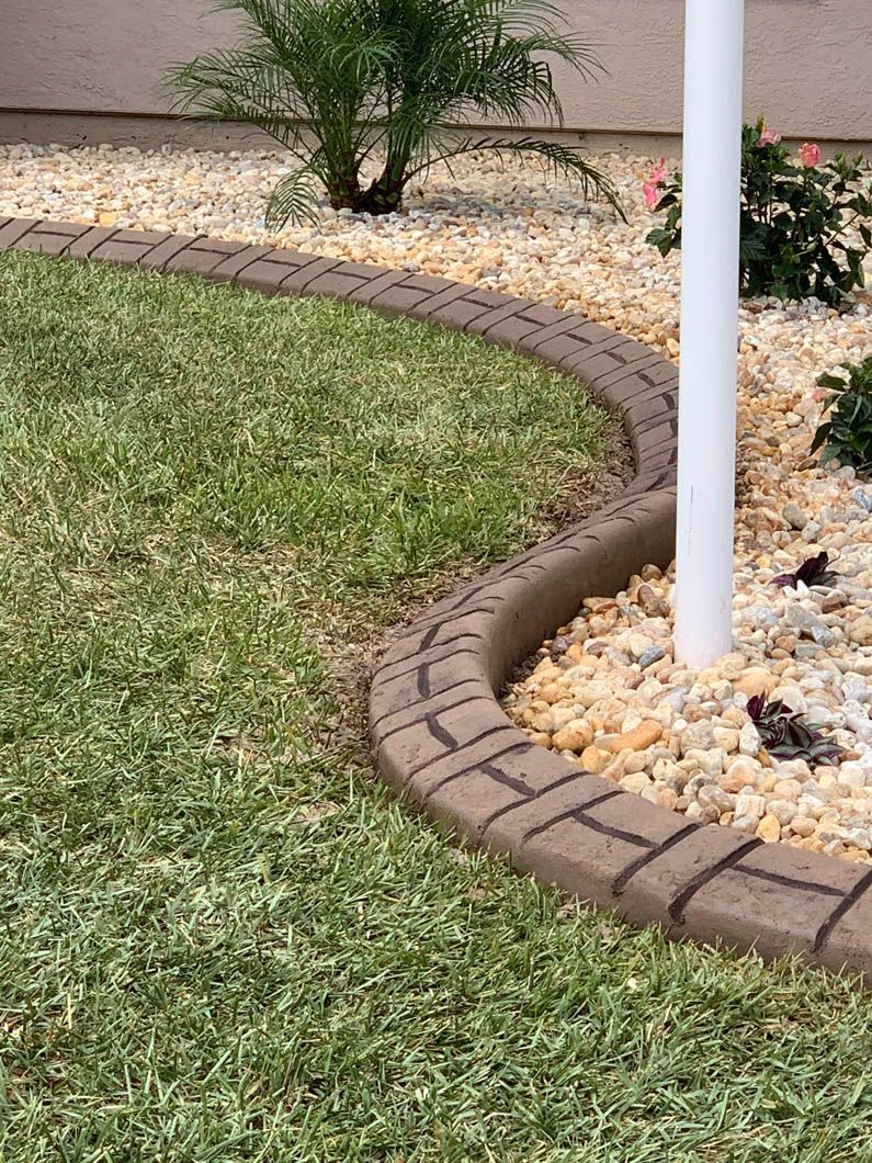 Curved brown concrete curbing borders green grass and tan rocks near a white post.
