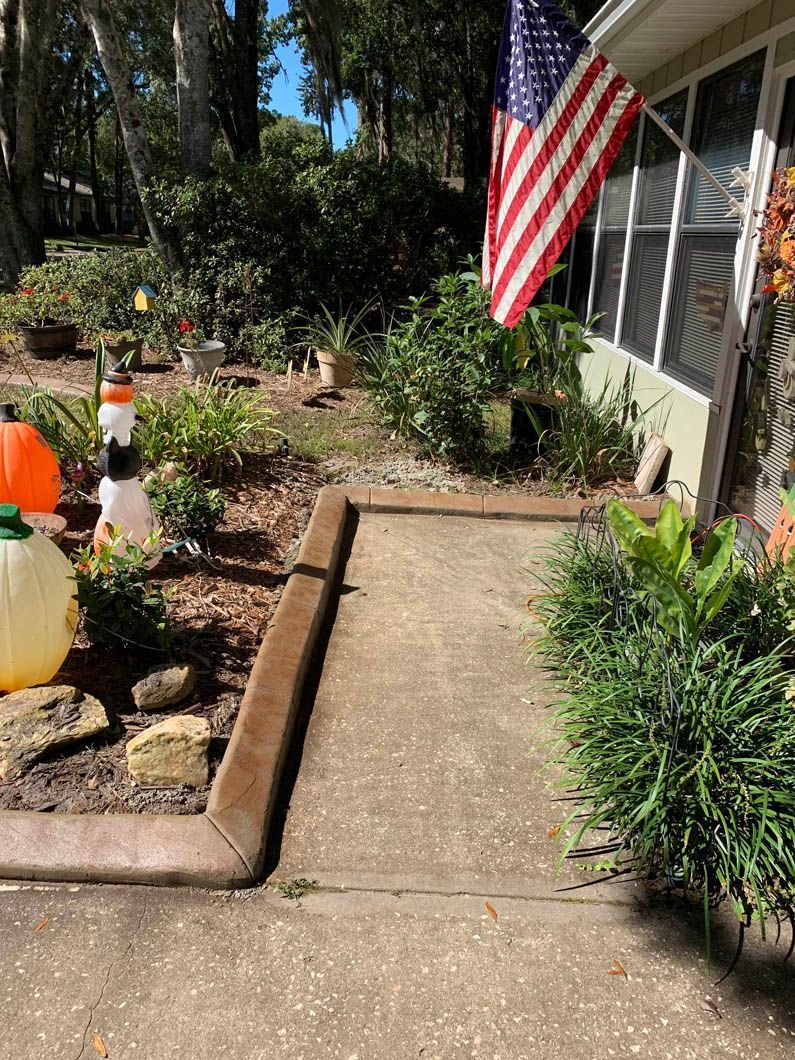 Pathway leading to a house, flanked by flowerbeds and American flag; daytime.