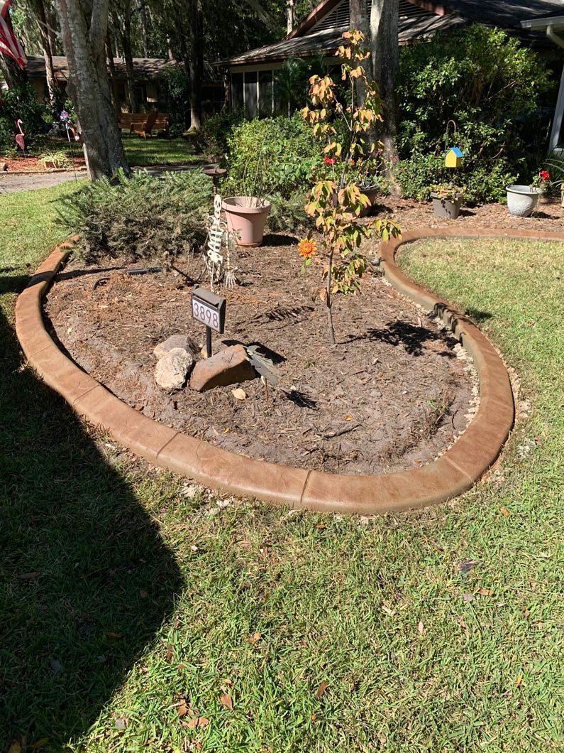 Brown concrete border surrounds a garden bed of mulch and plants, on a grassy lawn.