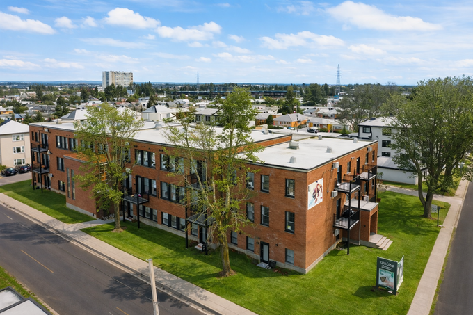Vue aérienne d'un immeuble d'appartements en briques, bordé de rues arborées par une journée ensoleillée.