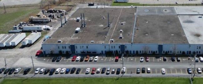 Vue aérienne d'un long bâtiment industriel avec des équipements sur le toit et des voitures garées le long de la façade.