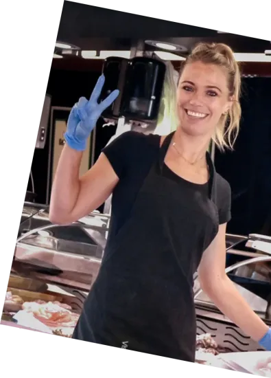 Een lachende cafémedewerker in een zwart schort en blauwe handschoenen houdt een display met zwarte kopjes omhoog.