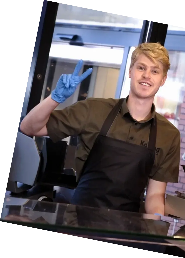 Een lachende cafémedewerker in schort en blauwe handschoenen maakt een vredesteken achter de toonbank.