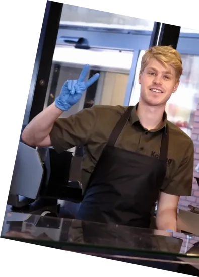 Een lachende barista in een zwart schort en blauwe handschoenen maakt een vredesteken achter de toonbank.