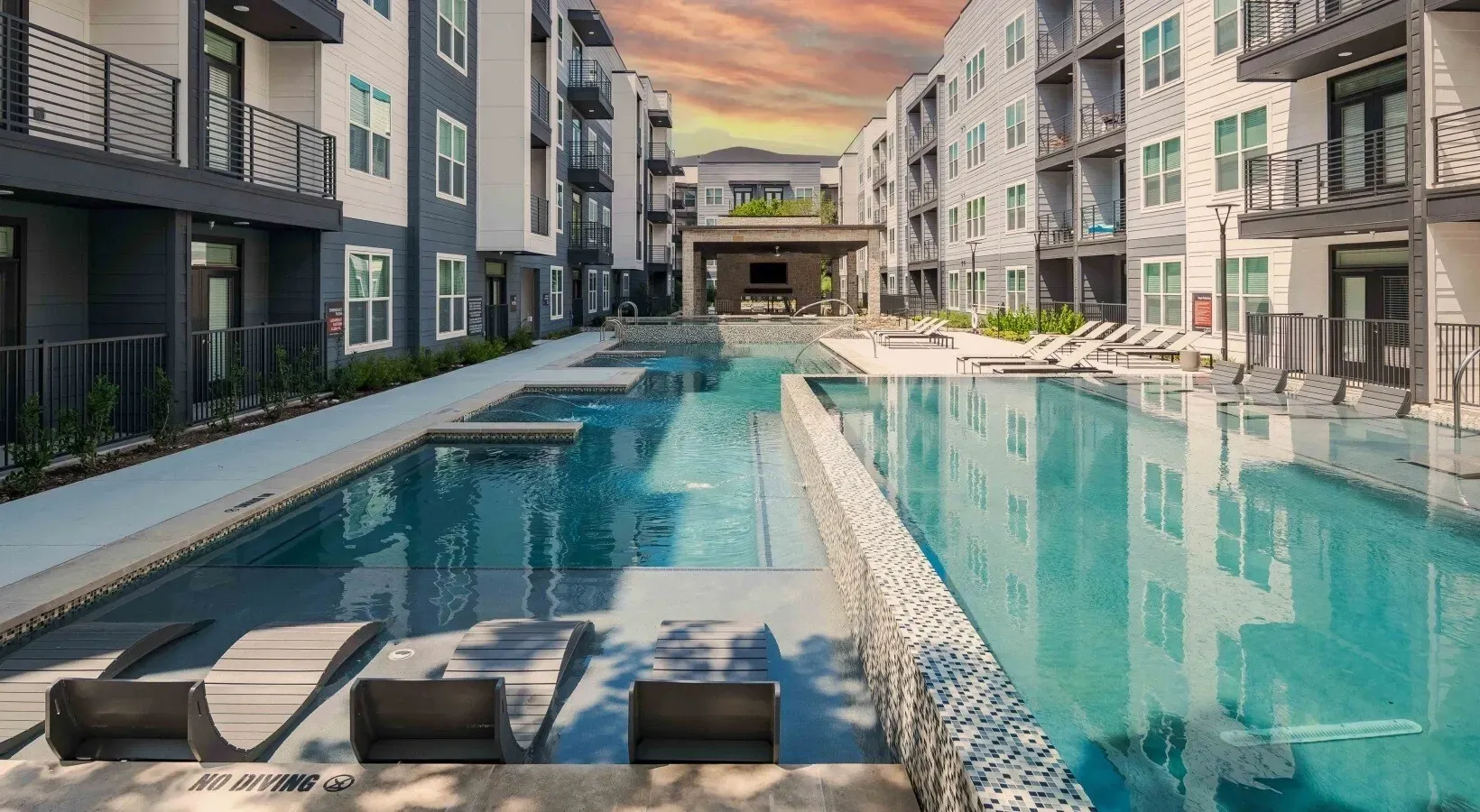 Modern apartment courtyard with a long turquoise pool at sunset, lined by lounge chairs and surrounding buildings