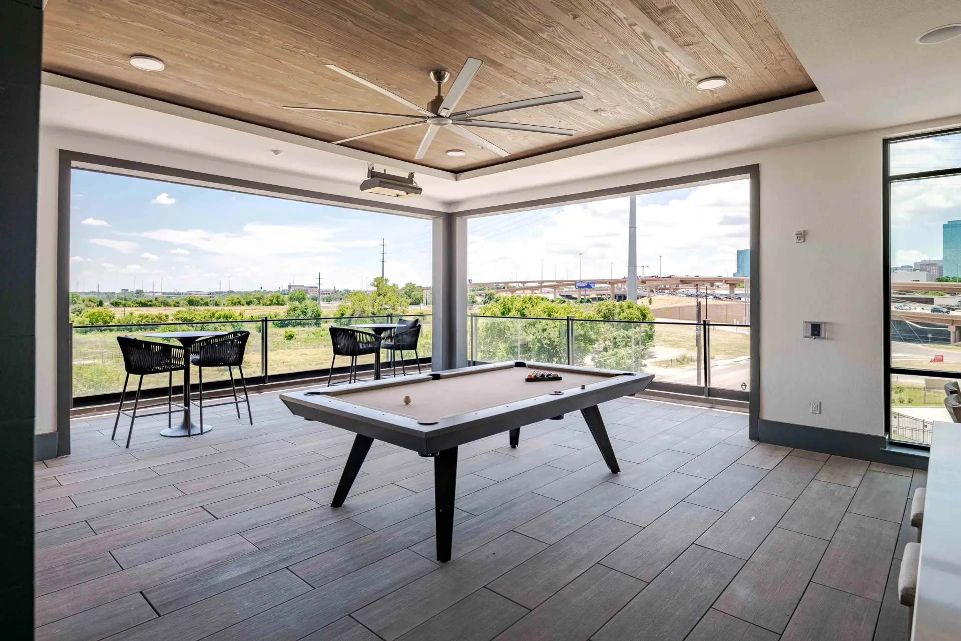 Modern patio with a pool table, chairs, and large open windows overlooking a green landscape.