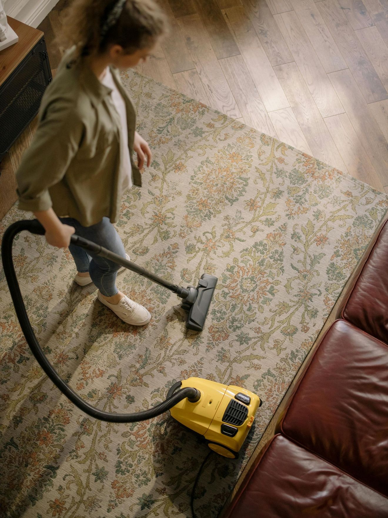 A person vacuums a patterned rug in a room with a brown leather couch.