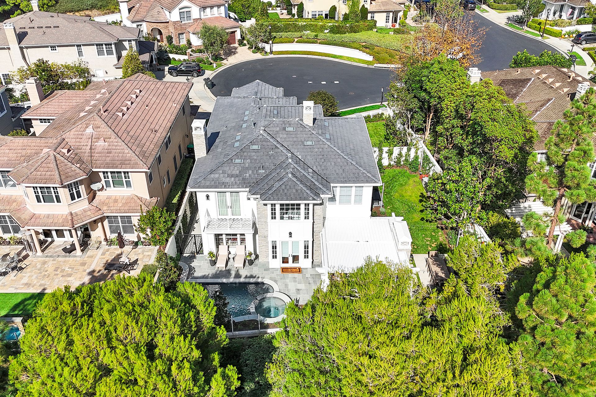 Aerial view of a large white house with a dark gray roof, surrounded by green trees and other houses.