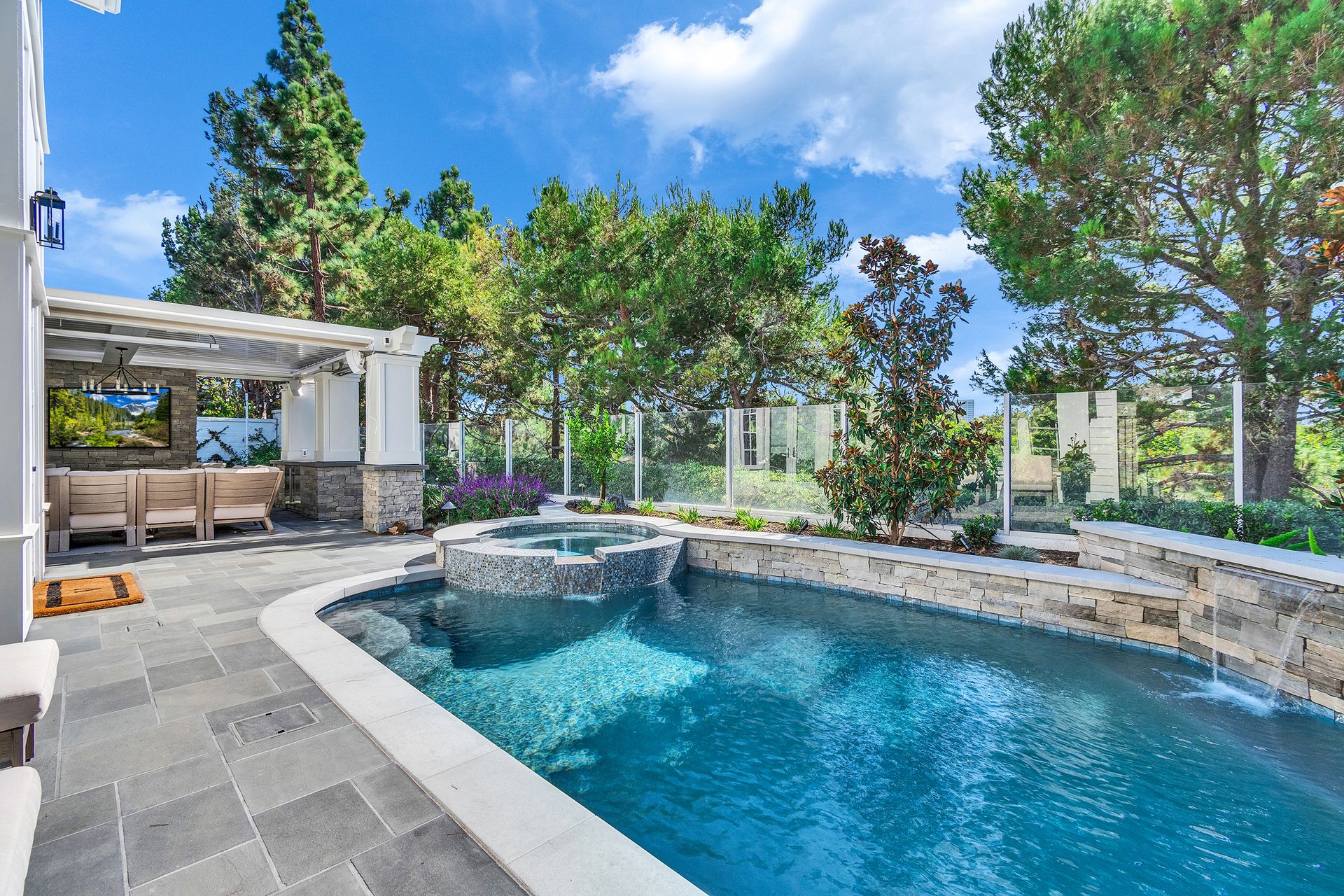 Backyard pool with spa, patio, and pergola, surrounded by trees under a blue sky.
