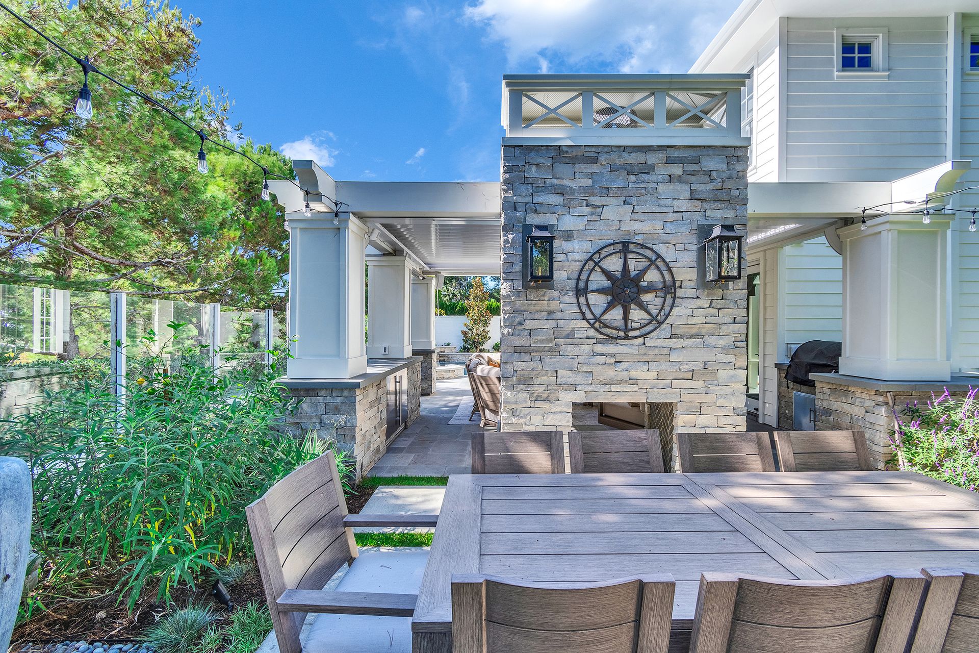 Patio with wooden furniture, stone fireplace, and white building exterior under blue sky.