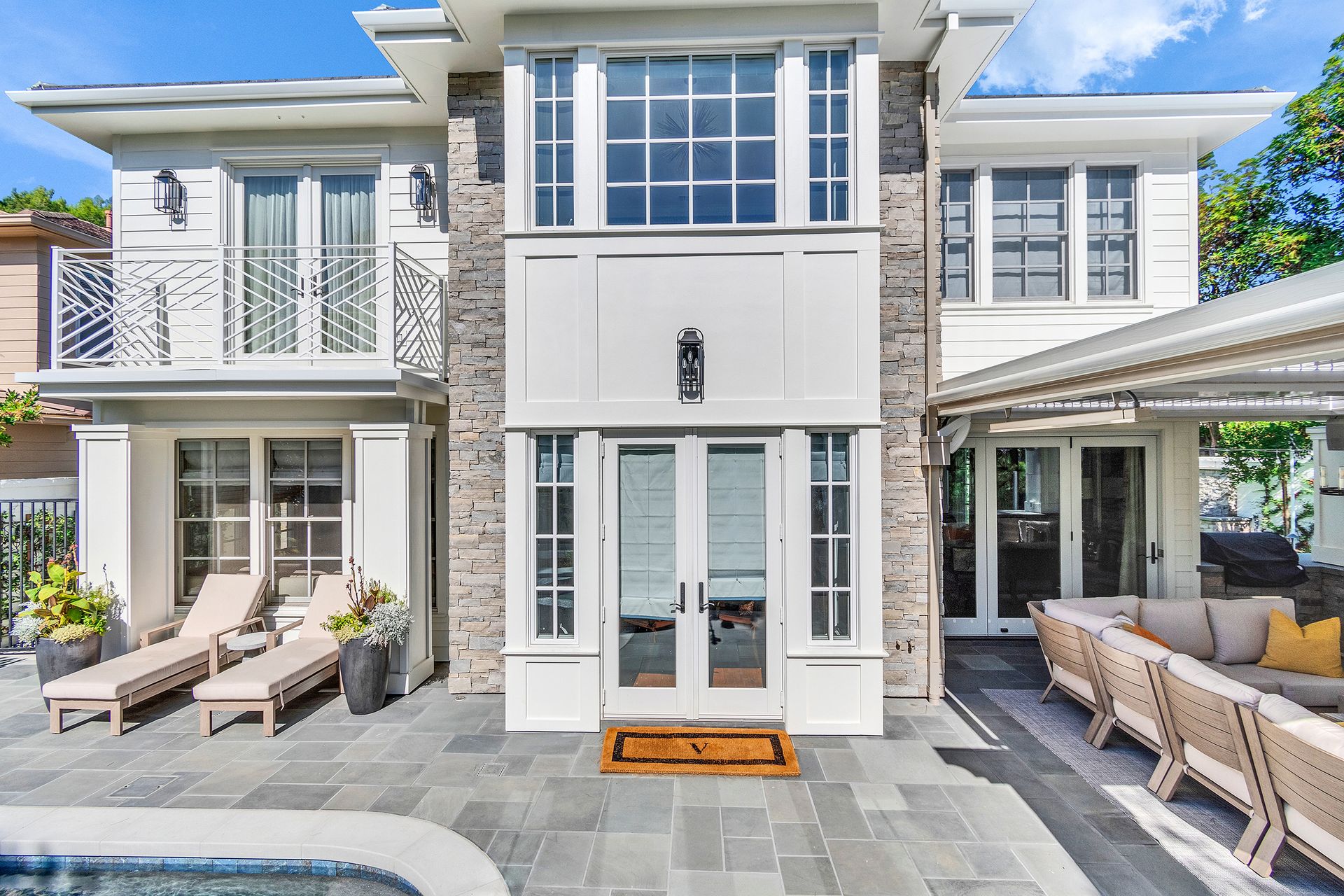 Two-story white house with stone accents, French doors, balcony, and outdoor patio with furniture, next to a pool.
