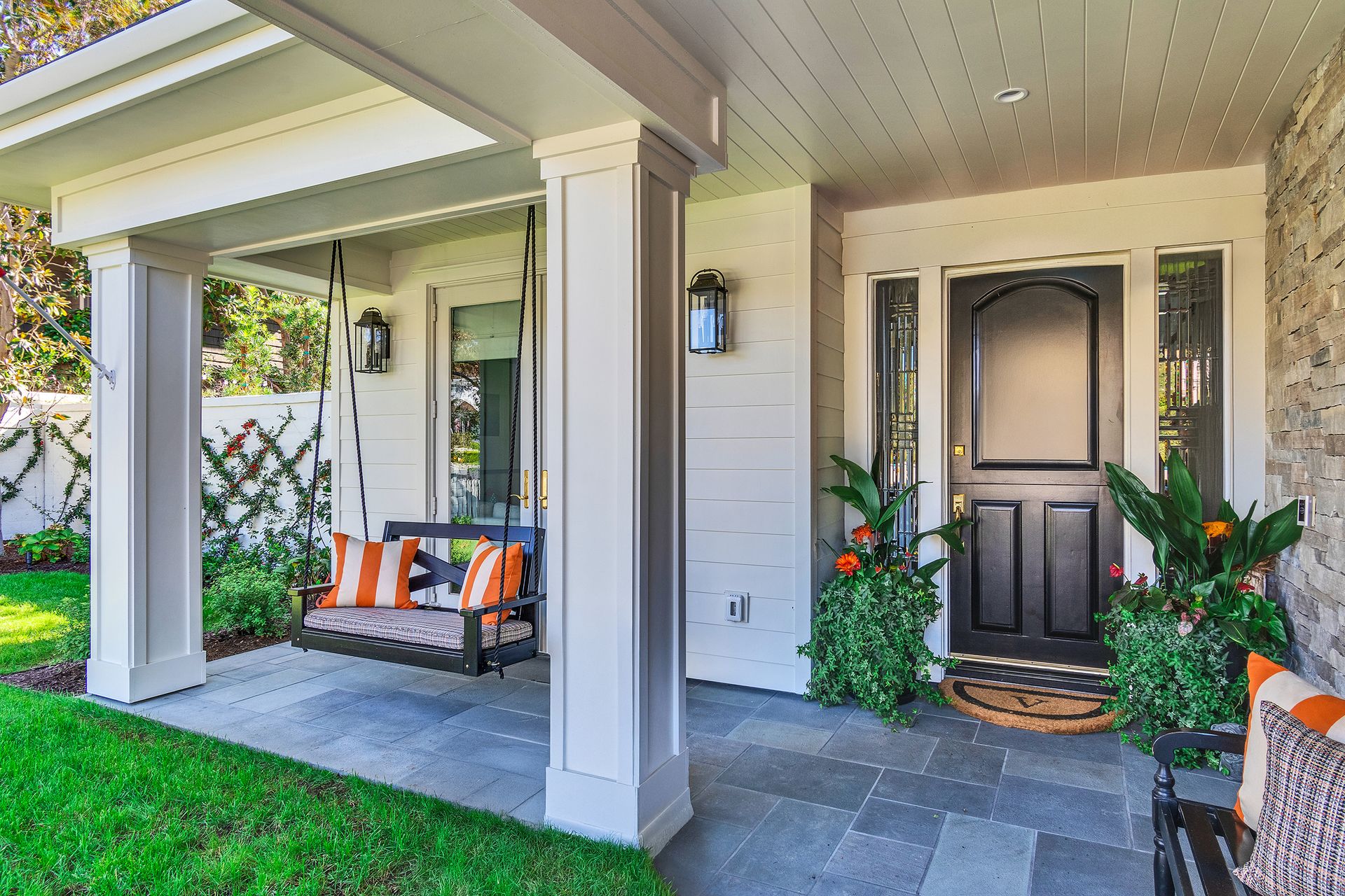 Front porch with swing, black door, and potted plants. Gray stone patio, white columns, green lawn.