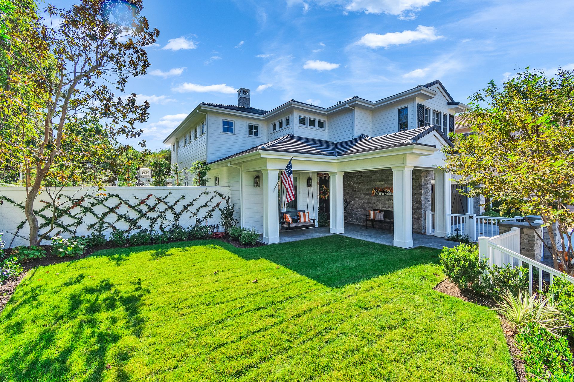 White house with a covered porch, lush green lawn, and climbing plants on a white fence under a blue sky.