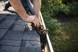 Hands clearing debris from a rain gutter on a shingled roof
