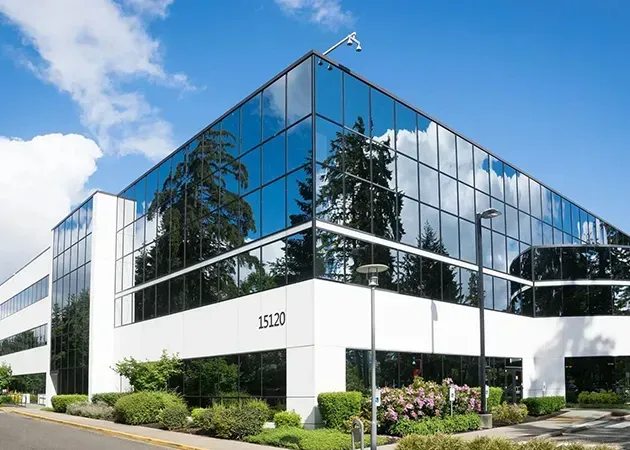 Modern glass office building with reflective windows, white facade, and landscaped entrance on a sunny day