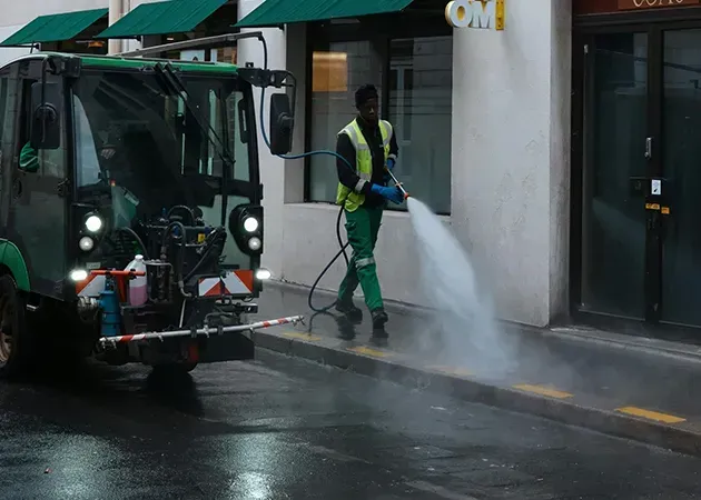 Street cleaner in a yellow vest washing a wet sidewalk beside a truck with a power hose.