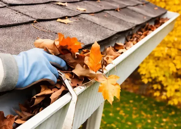 Gloved hand clearing autumn leaves from a roof gutter beside a house