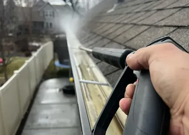 Hand holding a pressure washer wand spraying a house roof gutter from a ladder