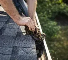 Hands clearing debris from a rain gutter on a shingled roof