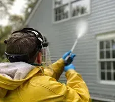Person in yellow raincoat pressure-washing a house exterior with a spray wand