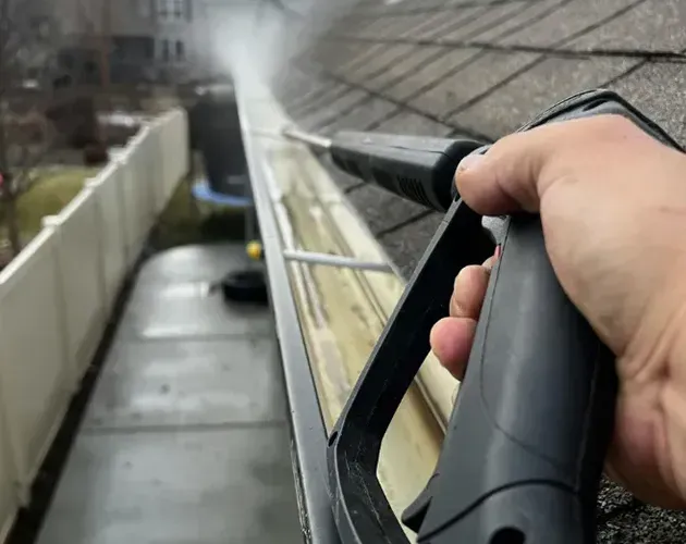 Hand spraying water from a roof gutter cleaning wand along a sloped shingle roof