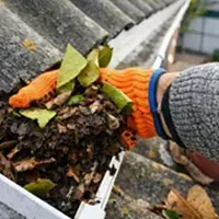 Orange-gloved hand empties a gutter scoop of wet leaves and debris beside a roof gutter