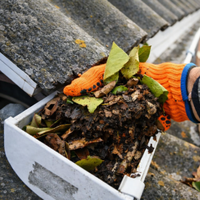 Orange-gloved hand empties a gutter scoop of wet leaves and debris beside a roof gutter