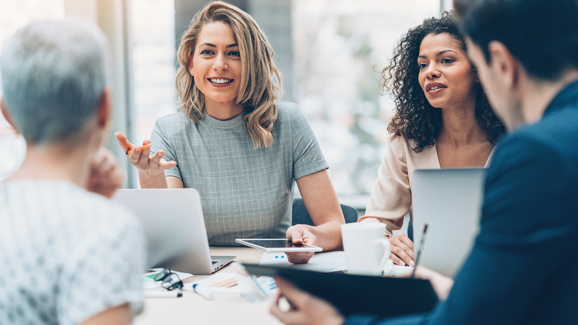 People at a meeting, talking. Blonde woman gesturing, smiling. Laptops, coffee mugs on table. Bright, modern office setting.