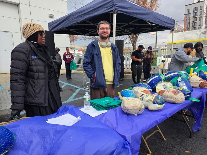 Two people stand behind a table with food items. Others stand nearby. Cloudy, outdoor setting.