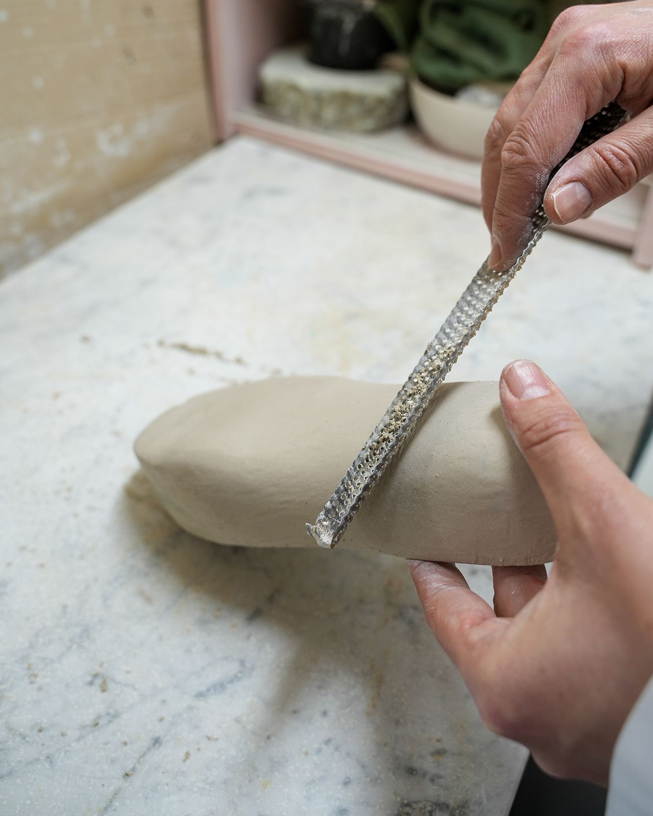 Mani che modellano l'argilla con una raspa metallica su una superficie bianca; ambiente di studio di ceramica.