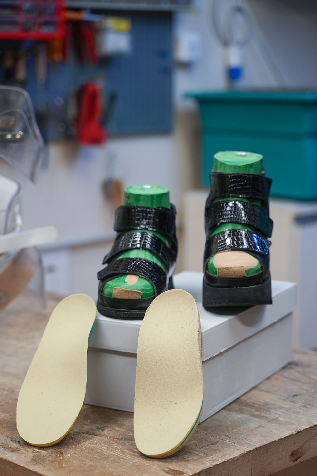 Due scarpe ortopediche nere su una scatola, con inserti. Sullo sfondo un'officina con attrezzi.