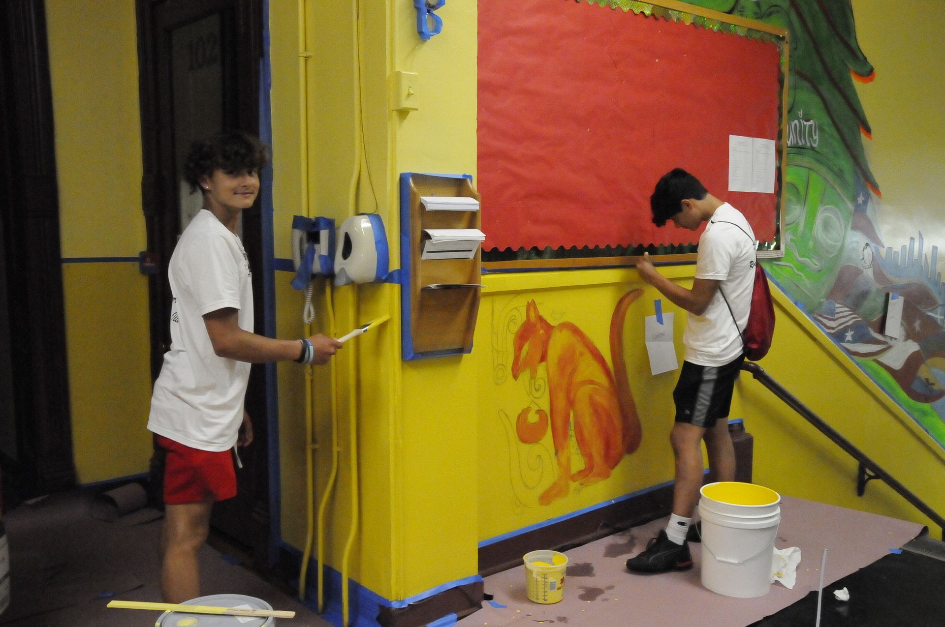 Two boys are painting a yellow wall with a squirrel on it