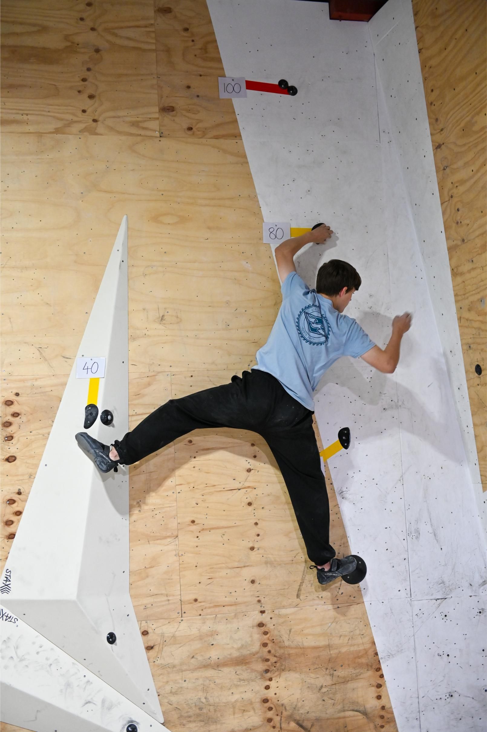 A person climbs a bouldering wall, arms stretched and legs wide, indoors.