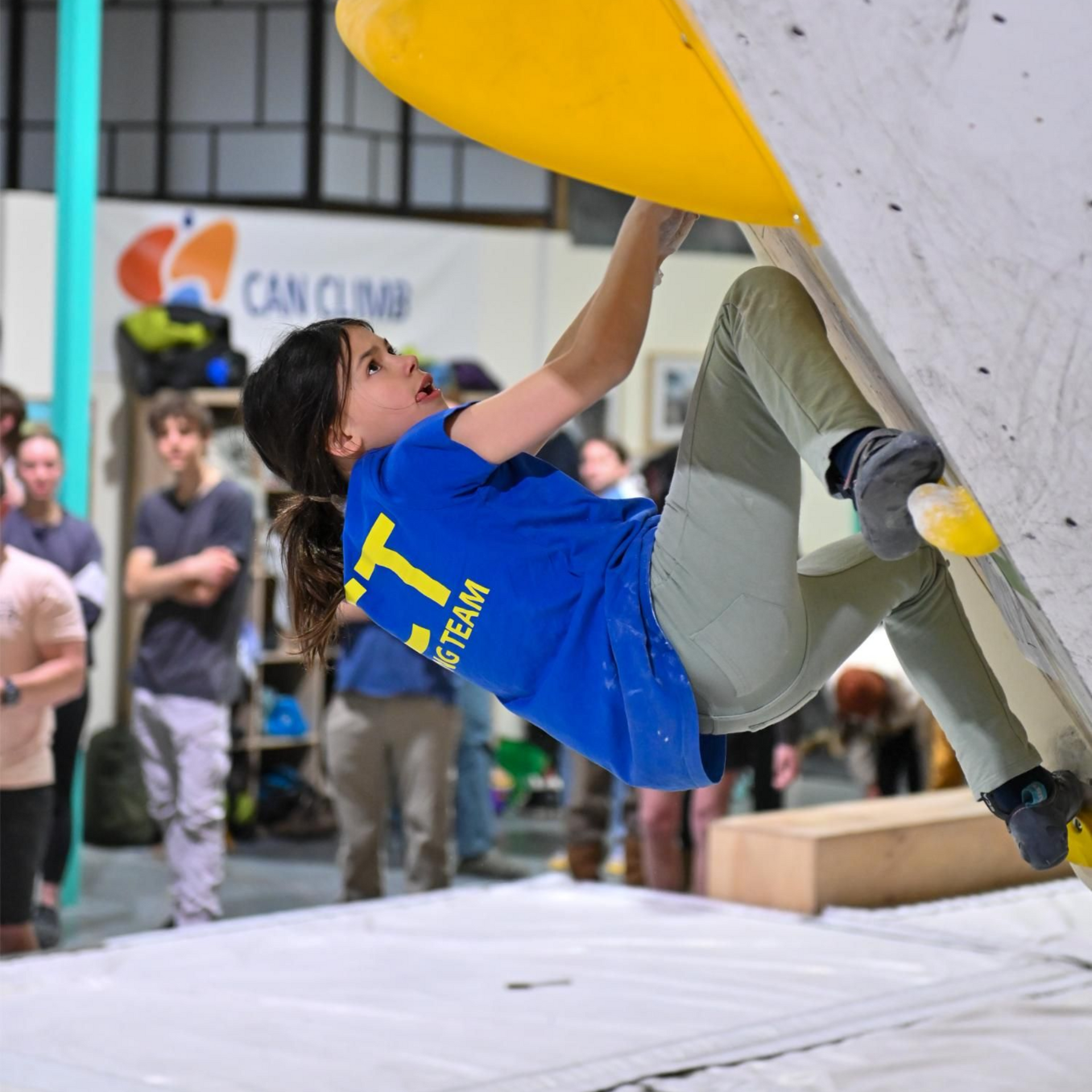A climber in blue shirt hangs from a yellow hold on a climbing wall, indoors.