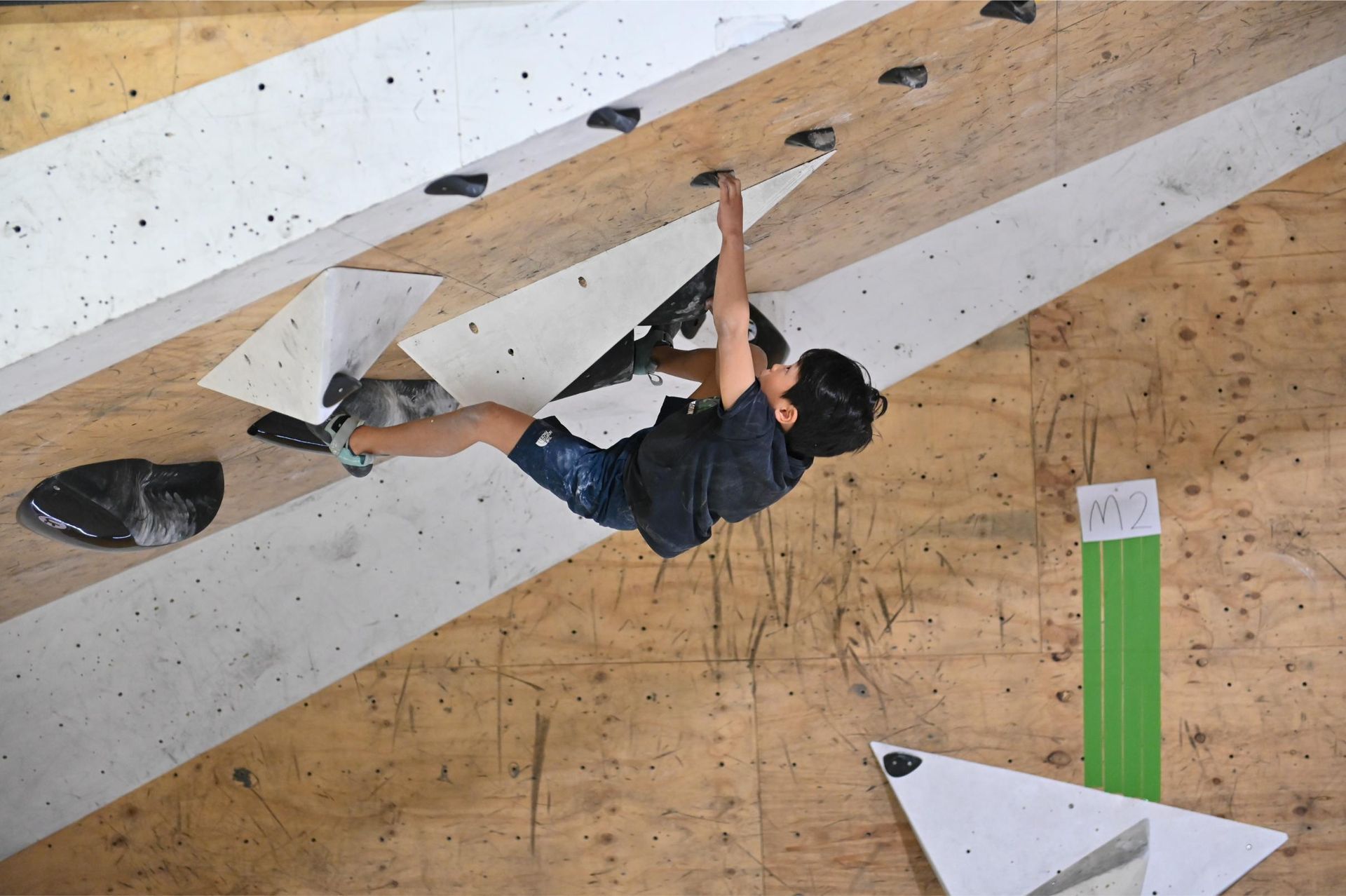 A person climbing an angled wall in a climbing gym, reaching for a handhold.