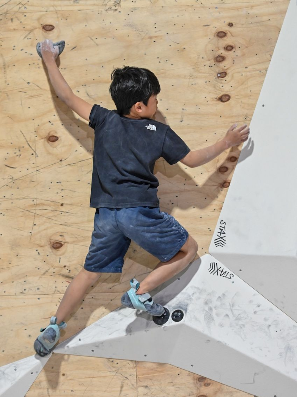 Young person climbing a wooden and white textured climbing wall, reaching with one arm.