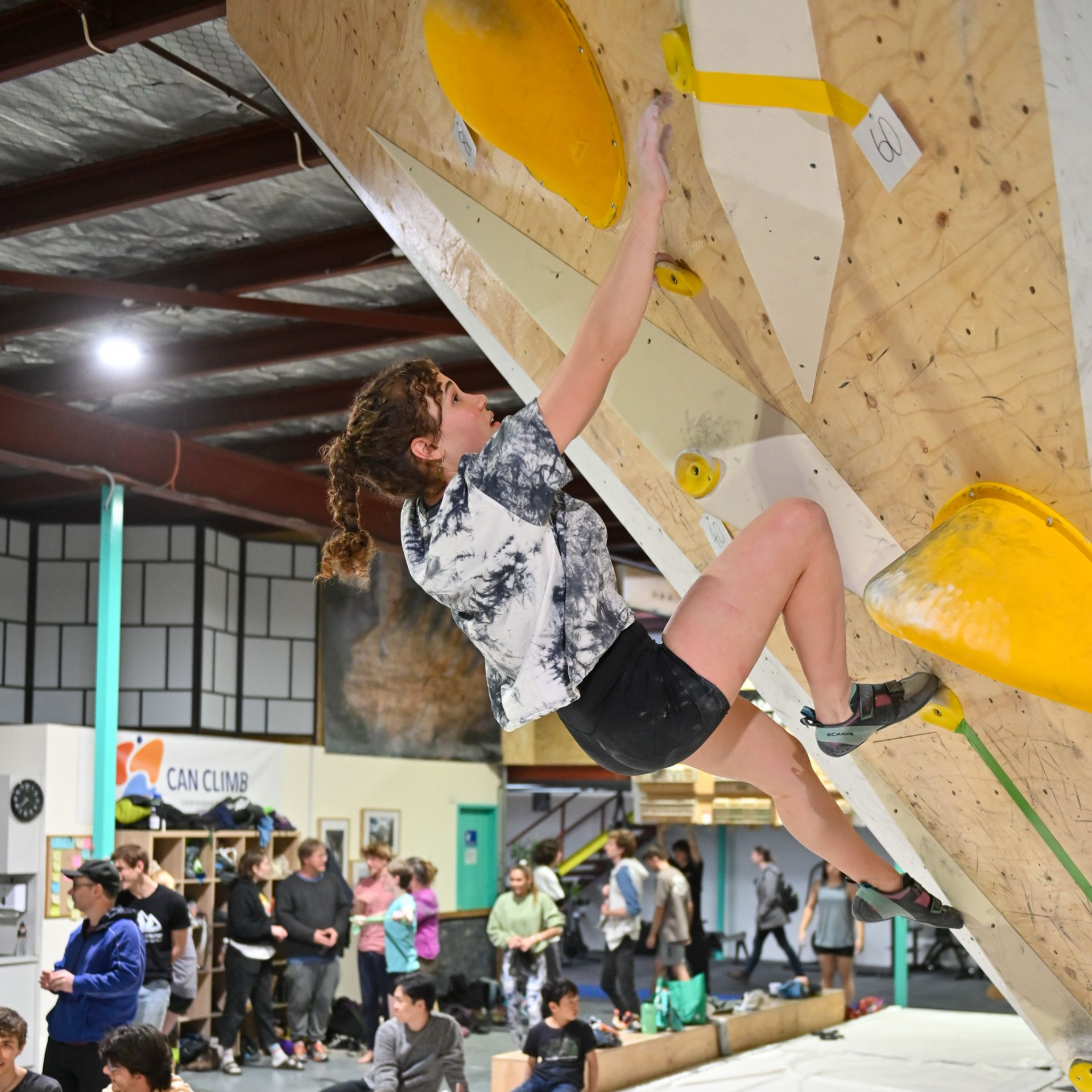 Person climbing an indoor rock wall, reaching for a yellow hold. Other people are in the background.