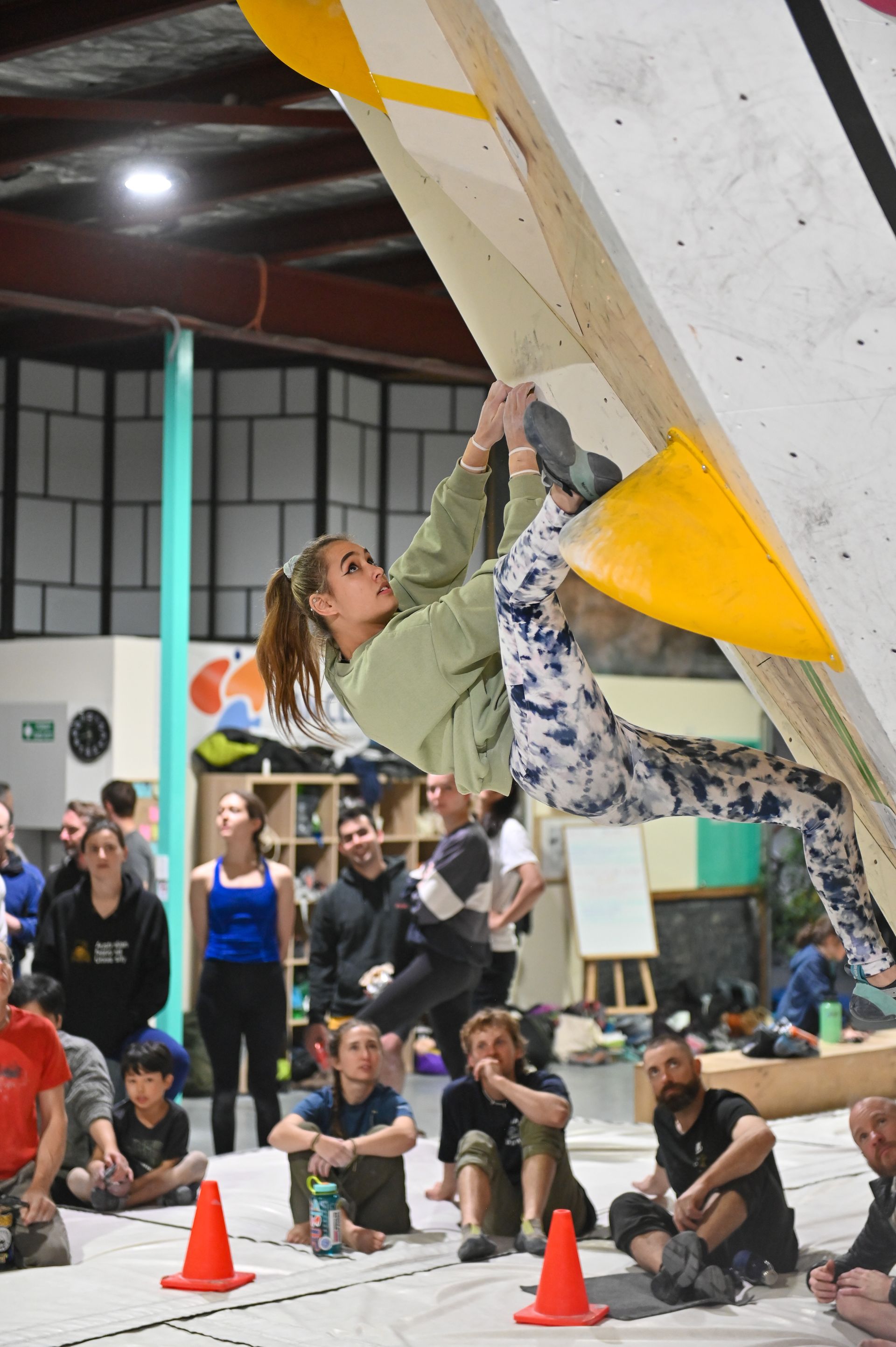 Woman climbing on an indoor bouldering wall, wearing a green top and patterned pants. Several people are watching.