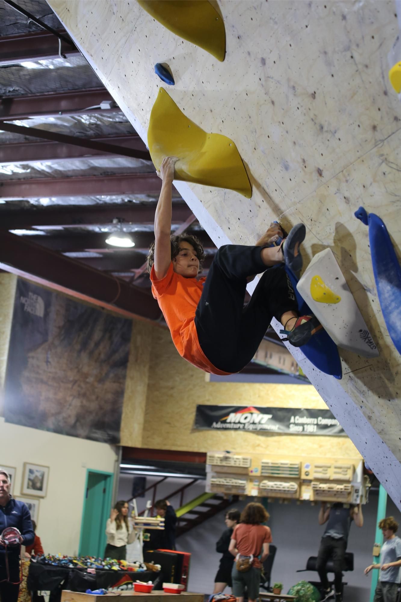 Climber in orange shirt grips yellow hold on an indoor climbing wall.
