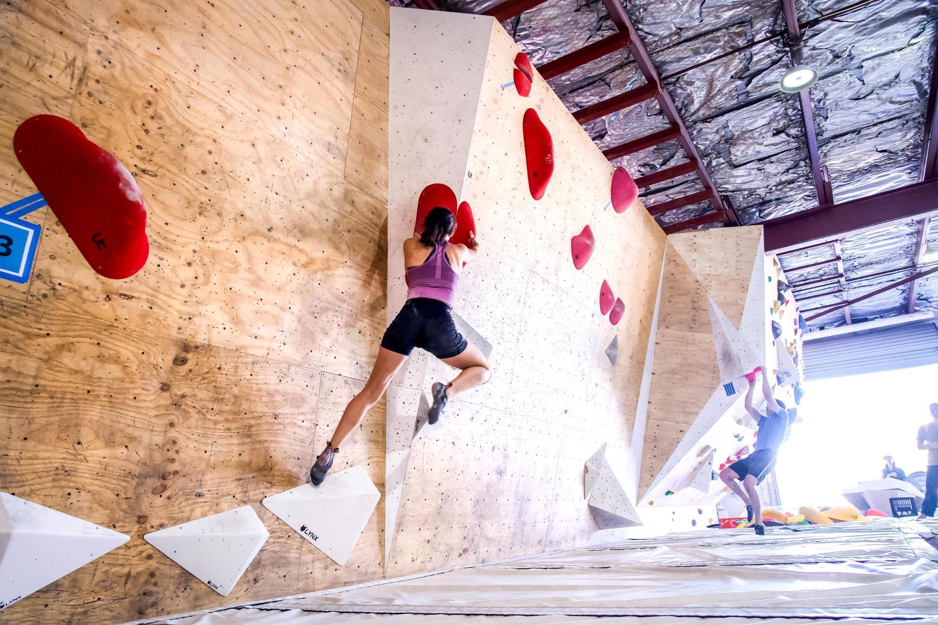 Woman climbing an indoor bouldering wall, reaching for red handholds, athletic wear, wood and white wall.