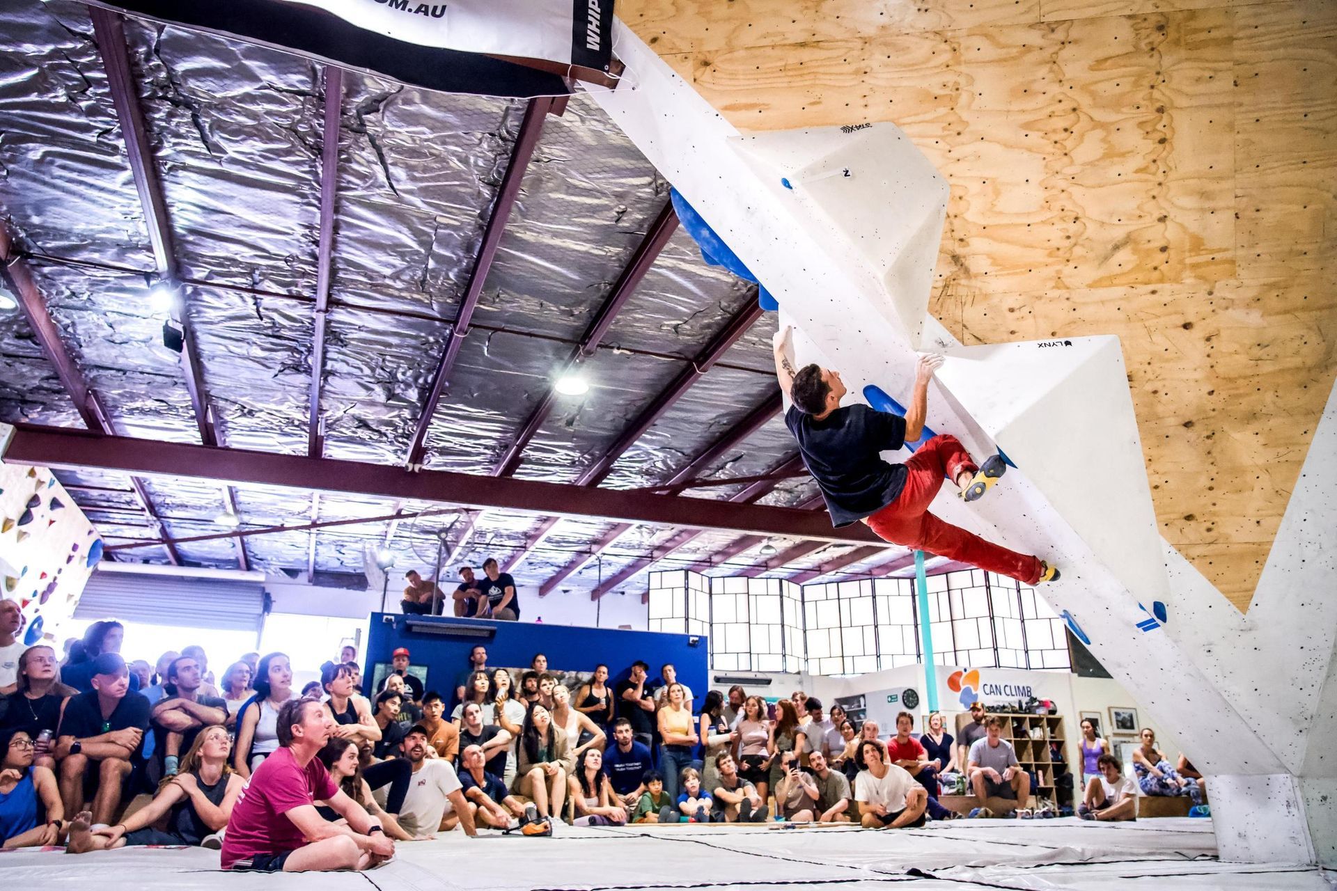 Climber in red pants on a white climbing wall, surrounded by a crowd in an indoor climbing gym.