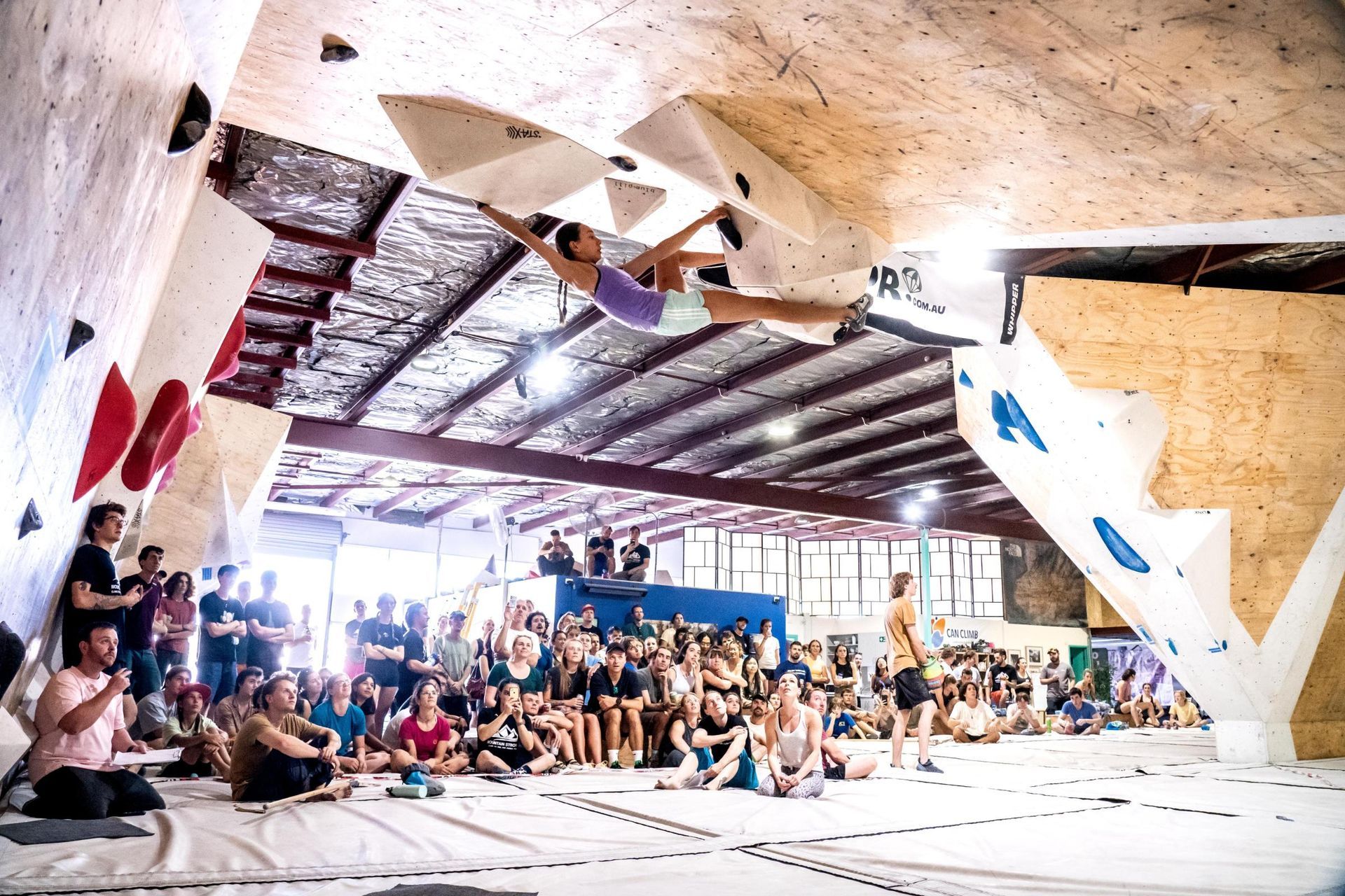 Woman bouldering, mid-air, in a climbing gym, cheered by a crowd.