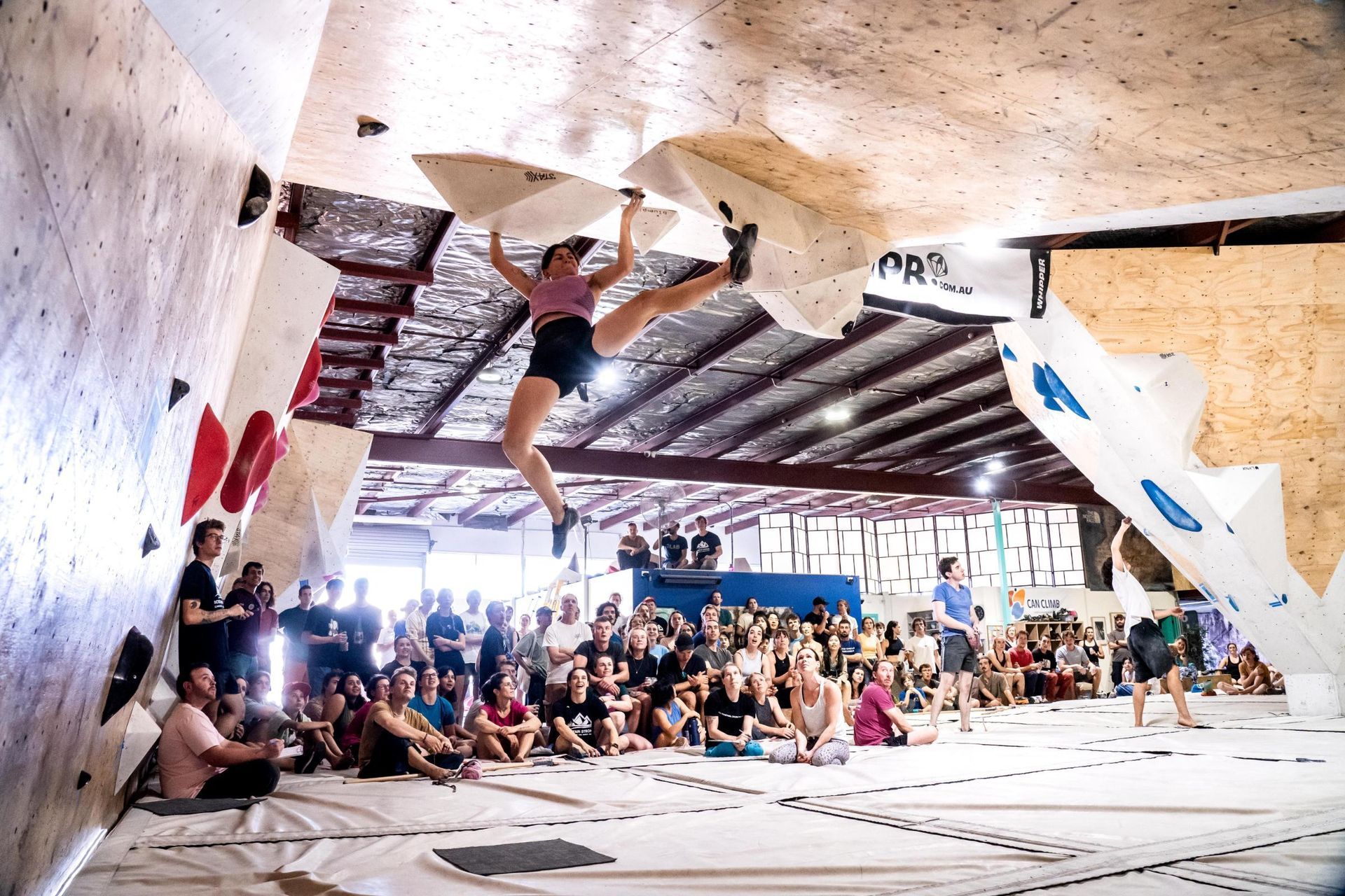 Woman bouldering indoors, legs split in mid-air, reaching for handholds on a textured wall. Many people watch.