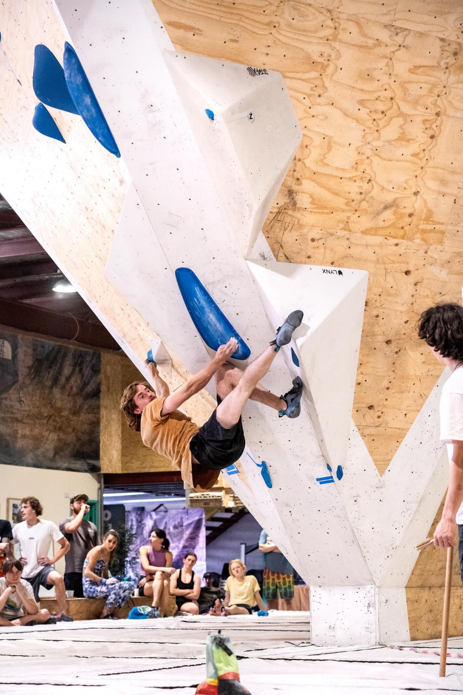 A person bouldering on a white climbing wall with blue holds; other people watch.