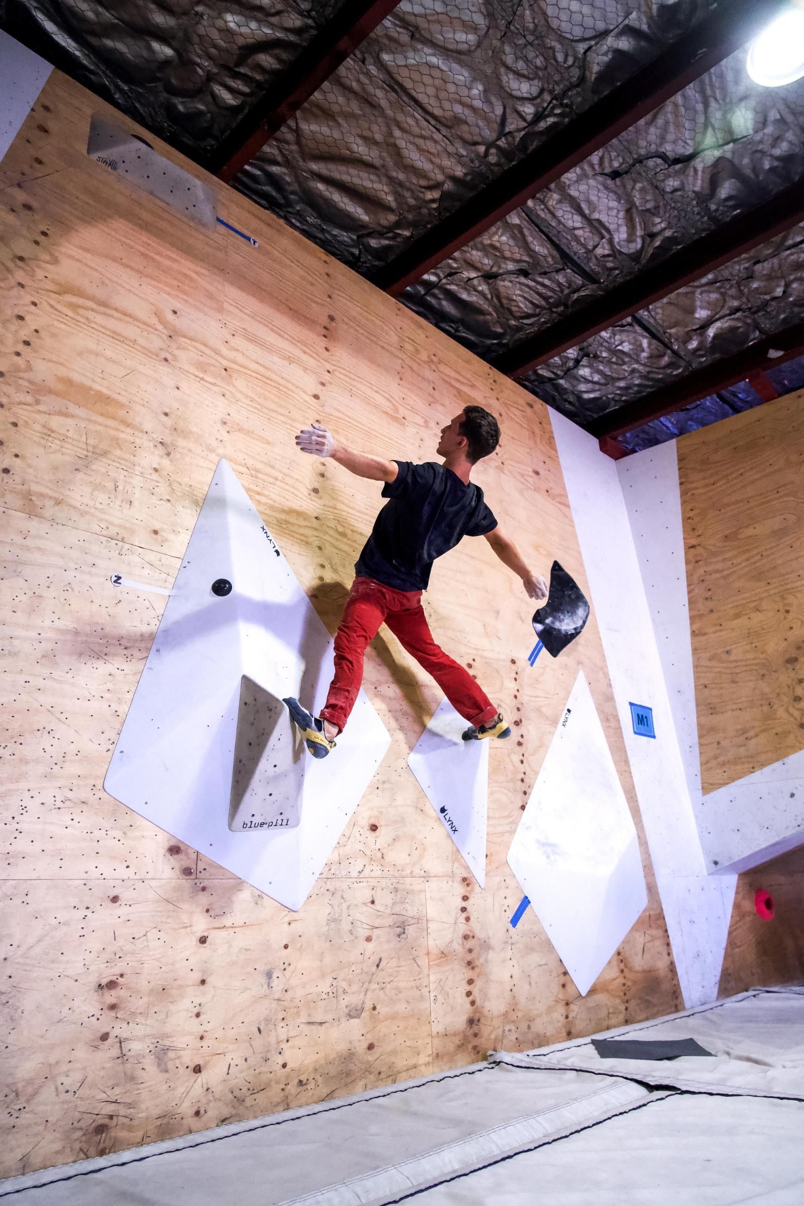 A person climbing a wall in a climbing gym. They are reaching for a hold, wearing red pants.