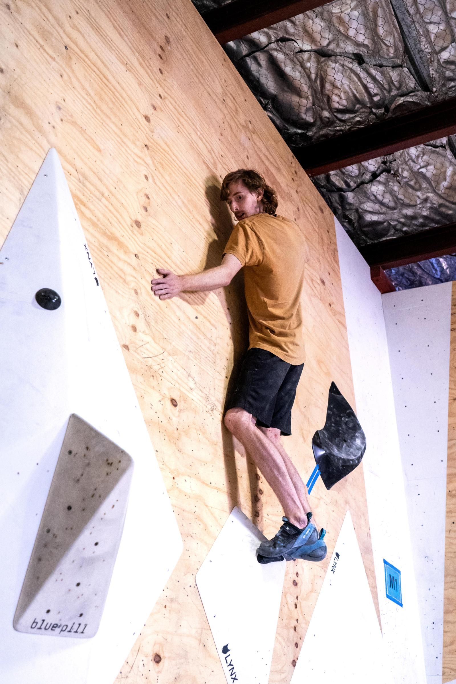Person climbing a wooden climbing wall, wearing shorts and shirt.