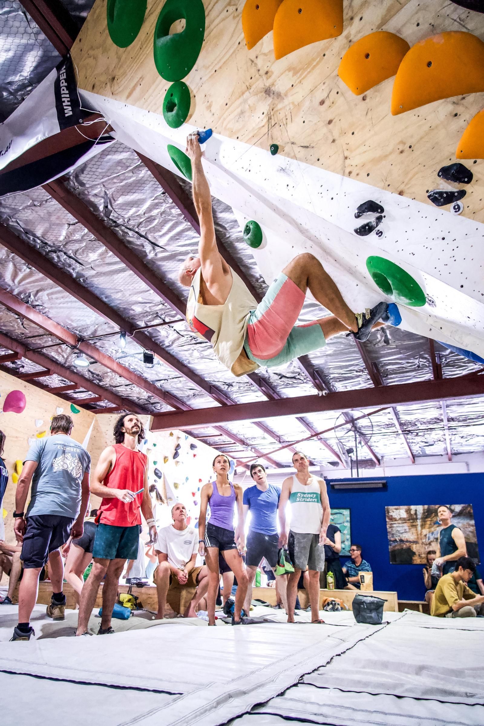 Rock climber on an overhanging wall with onlookers. Indoor climbing gym with colorful holds.