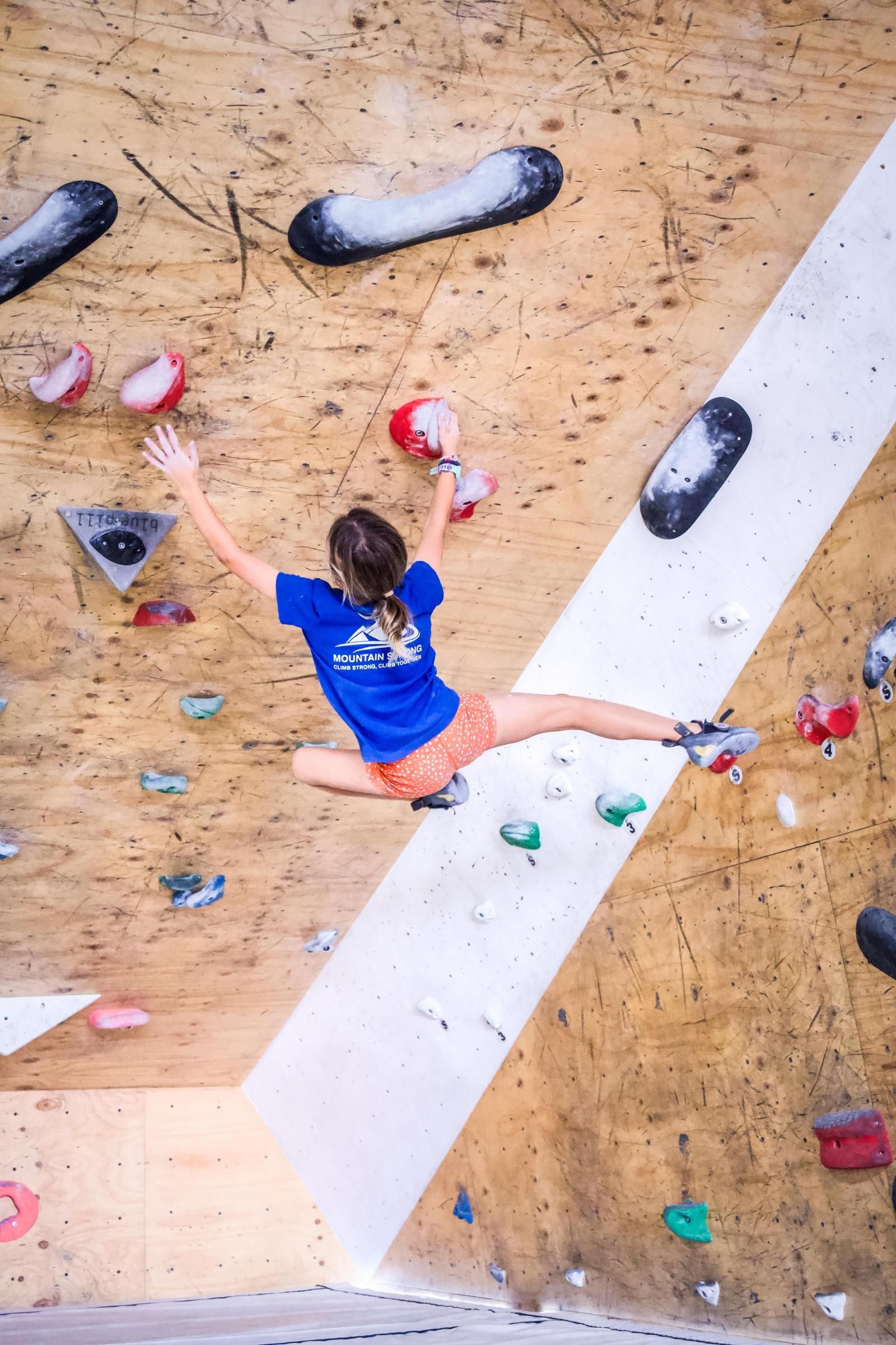 Person rock climbing on an angled wall, legs split. Wearing blue shirt and orange shorts, arms outstretched.